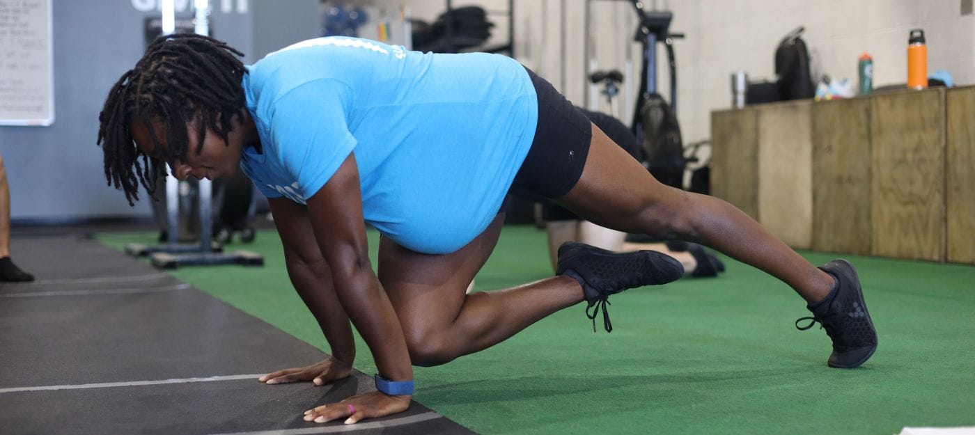 A pregnant woman with dark hair and a blue shirt performs a Scorpion Reach in a gym.